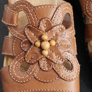 May include: A brown leather slide sandal featuring a decorative cutout floral pattern on the upper strap. The center of the flower is adorned with four small, round, light tan beads. The sandal includes light-colored contrast stitching throughout.