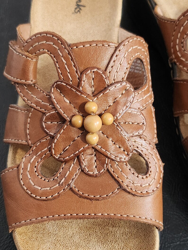 May include: A brown leather slide sandal featuring a decorative cutout floral pattern on the upper strap. The center of the flower is adorned with four small, round, light tan beads. The sandal includes light-colored contrast stitching throughout.