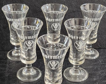 May include: Six clear glass shot glasses with a white deer logo and the text "Jägermeister" etched on the side. The glasses have a flared top and a short stem.