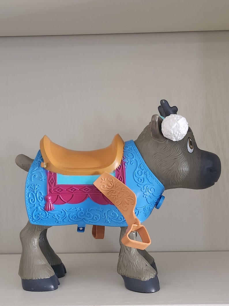 Pu&ograve; includere: Un giocattolo di renna in plastica con una sella. La renna &egrave; grigia con corna e zoccoli neri. Ha una coperta blu con accenti rosa e oro. Un pompon bianco &egrave; sulla sua testa.
