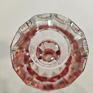 Op de afbeelding: Een helder, facetgeslepen glazen object met een roze tint. Het object heeft een cirkelvormige basis en een geschulpte rand. Het ontwerp bevat geometrische vormen en patronen, waardoor licht- en schaduweffecten ontstaan. Het object lijkt een decoratief item te zijn.