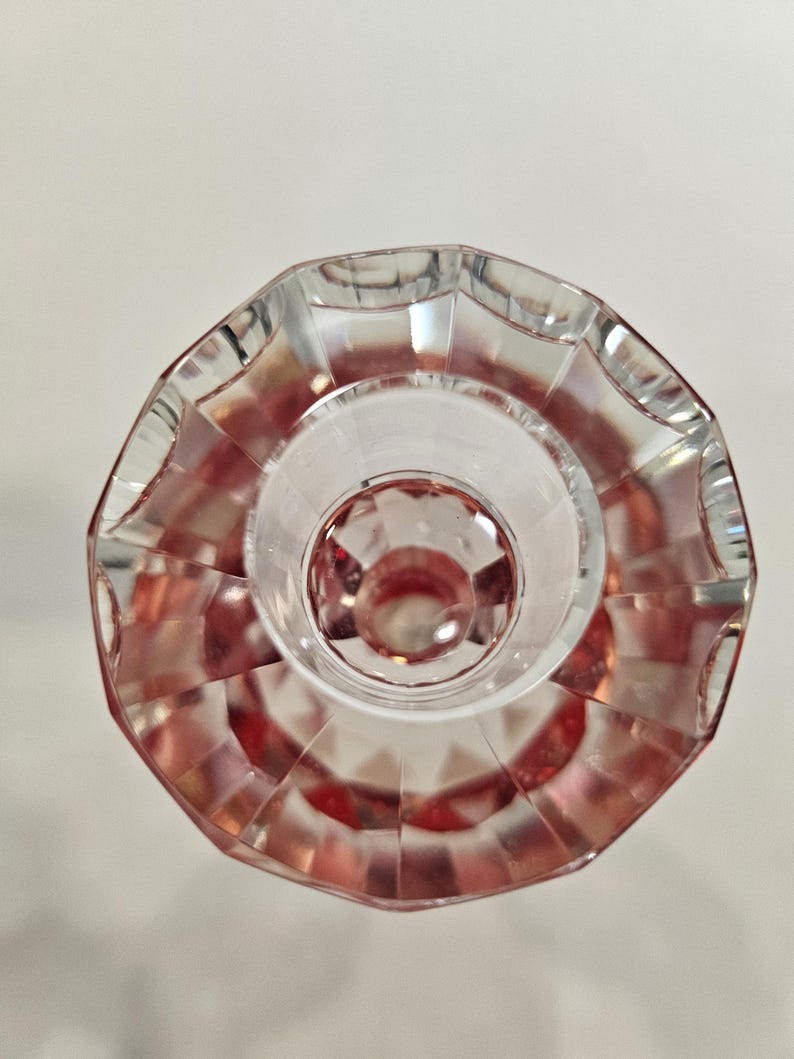 Op de afbeelding: Een helder, facetgeslepen glazen object met een roze tint. Het object heeft een cirkelvormige basis en een geschulpte rand. Het ontwerp bevat geometrische vormen en patronen, waardoor licht- en schaduweffecten ontstaan. Het object lijkt een decoratief item te zijn.