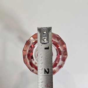 Op de afbeelding: Een heldere, facetgeslepen, rode glazen flesstopper met een zilveren meetlint ernaast. De stopper is ongeveer 5 cm hoog.