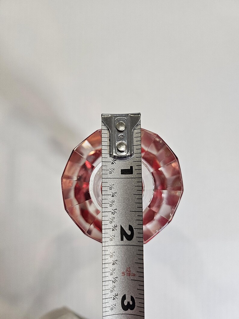 Op de afbeelding: Een heldere, facetgeslepen, rode glazen flesstopper met een zilveren meetlint ernaast. De stopper is ongeveer 5 cm hoog.