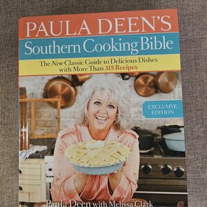 May include: A cookbook titled "Paula Deen's Southern Cooking Bible: The New Classic Guide to Delicious Dishes with More Than 315 Recipes" by Paula Deen with Melissa Clark. The cover features a photo of Paula Deen holding a pie in a kitchen setting.