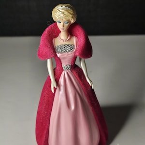 May include: A pink and red plastic doll wearing a crown and a pink dress with a red velvet cape.