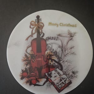 May include: A white round ceramic plate with a Christmas design. The plate features a red violin, a gold bow, a gold trumpet, a book with the word "Bible" on the cover, and the words "Merry Christmas" in gold lettering.