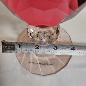 Op de afbeelding: Een facetrijk, robijnrood decoratief object met een heldere, kristallen steel en voet. De voet heeft een diameter van ongeveer 7.6 cm. Het woord "Anthropologie" is op de voet gedrukt.