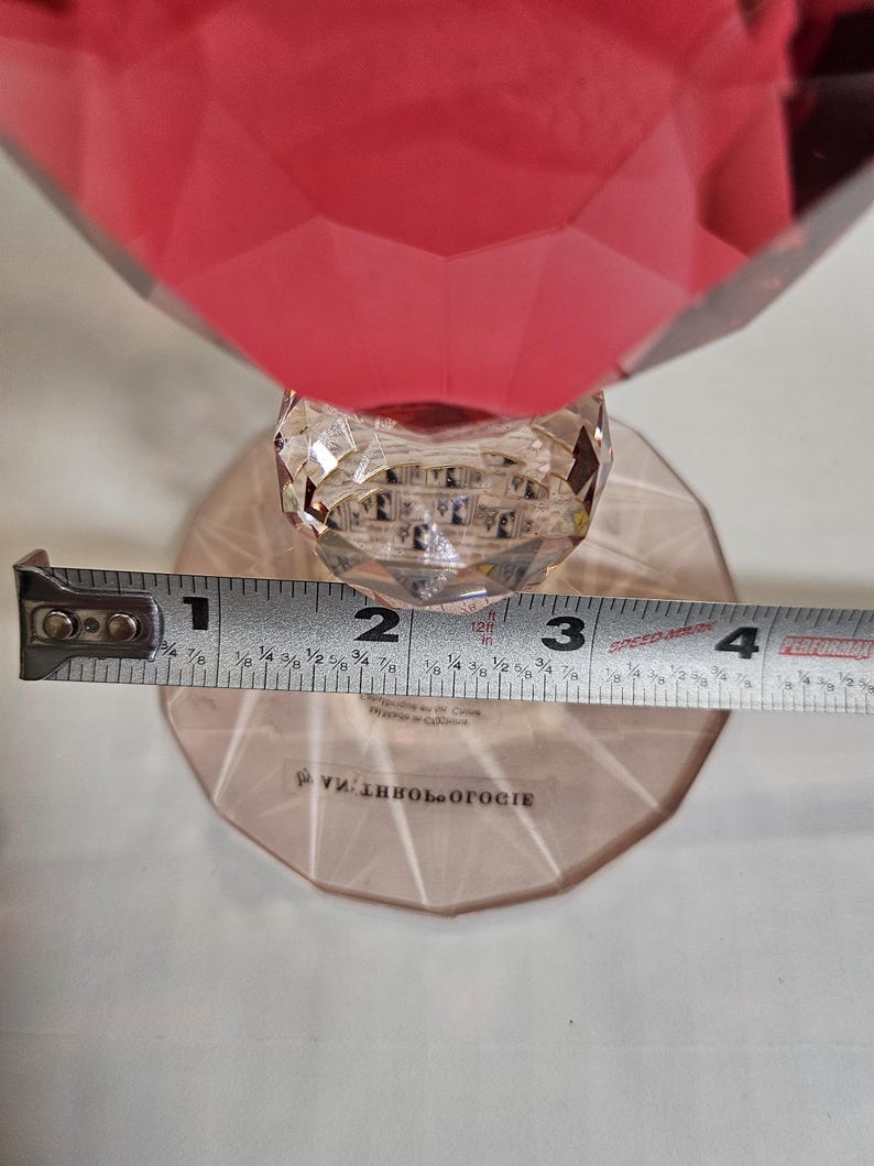 Op de afbeelding: Een facetrijk, robijnrood decoratief object met een heldere, kristallen steel en voet. De voet heeft een diameter van ongeveer 7.6 cm. Het woord "Anthropologie" is op de voet gedrukt.