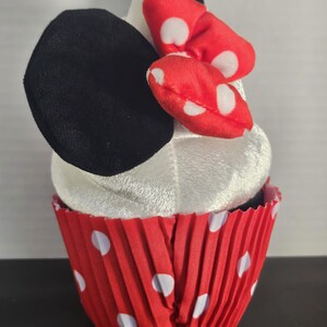 May include: A plush cupcake toy with a white top, black ears, and a red bow with white polka dots. The cupcake sits in a red paper liner with white polka dots, resembling a cartoon character.