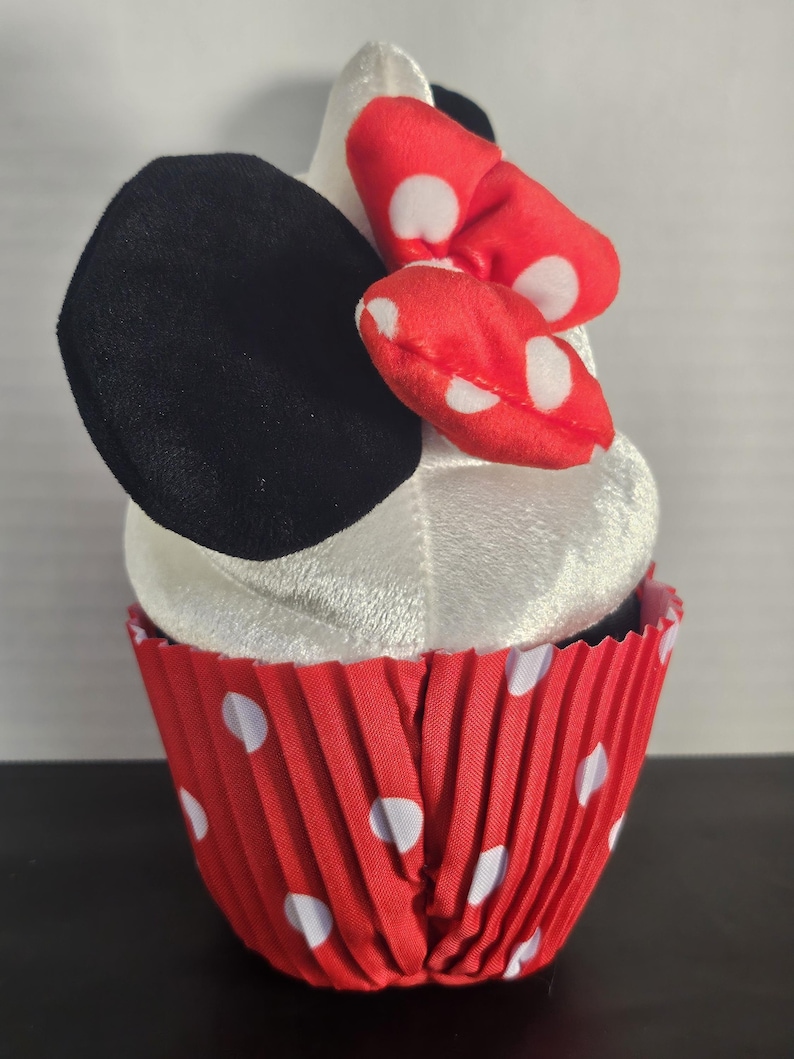 May include: A plush cupcake toy with a white top, black ears, and a red bow with white polka dots. The cupcake sits in a red paper liner with white polka dots, resembling a cartoon character.