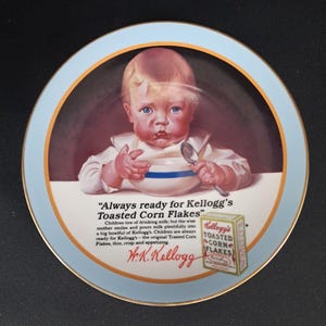 May include: A vintage plate featuring an illustration of a baby with a bowl and spoon, surrounded by text that reads "Always ready for Kellogg's Toasted Corn Flakes." The plate has a light blue border with gold trim.