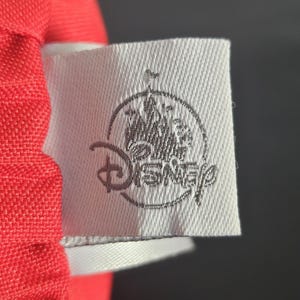 May include: Close-up of a white fabric tag with the Disney logo in gray. The tag is attached to a red fabric. The Disney logo features a castle and the word "Disney".