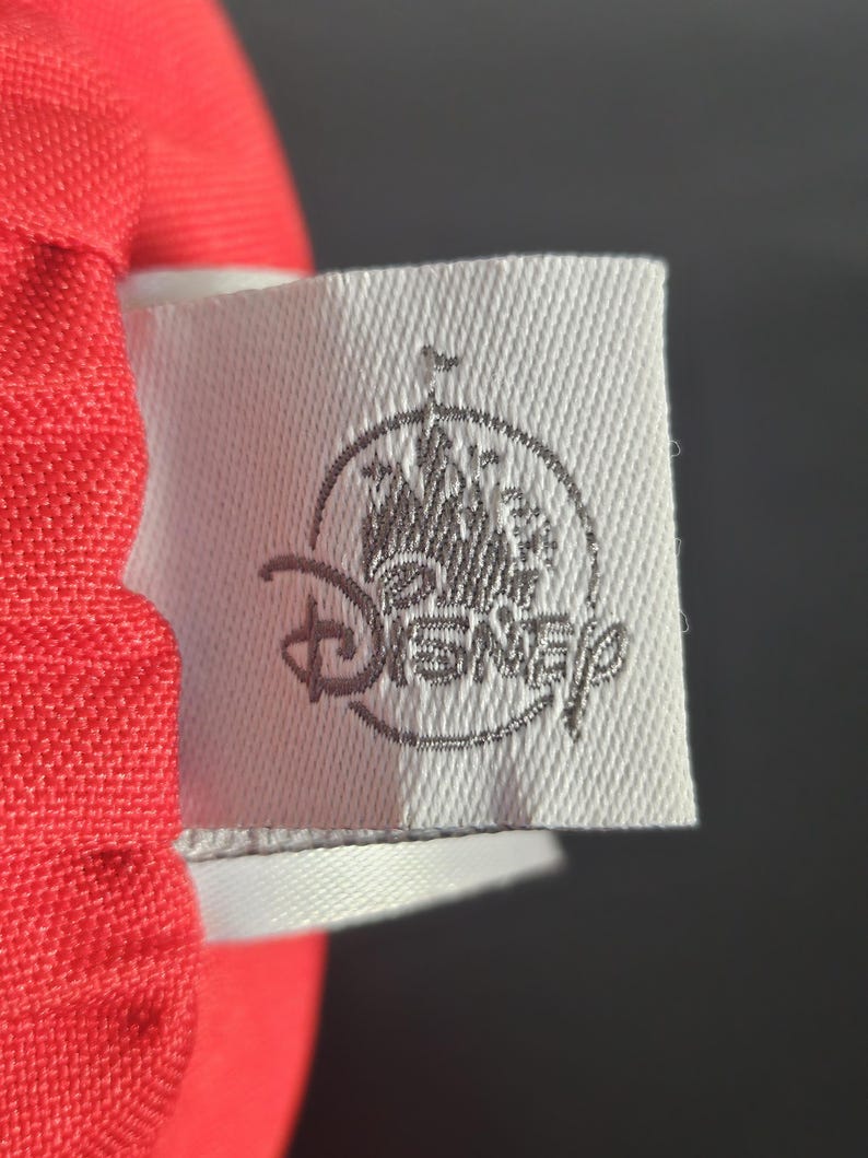 May include: Close-up of a white fabric tag with the Disney logo in gray. The tag is attached to a red fabric. The Disney logo features a castle and the word "Disney".