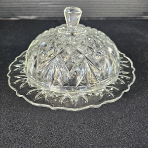 May include: A clear glass butter dish with a domed lid and a scalloped edge base. The lid has a diamond pattern and a small, faceted knob. The dish is designed to hold butter and is made of clear glass.