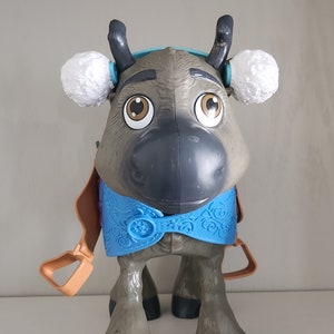 Pu&ograve; includere: Un giocattolo di plastica a forma di renna con grandi occhi marroni, corna grigie e paraorecchie bianche. La renna ha un gilet blu con una chiusura a fiocco di neve e staffe color cuoio. Il giocattolo &egrave; progettato per assomigliare a un personaggio del film Frozen.