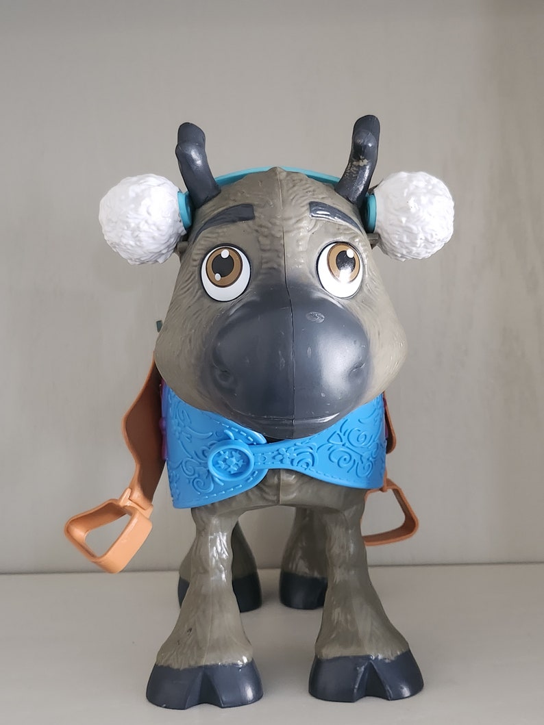 Pu&ograve; includere: Un giocattolo di plastica a forma di renna con grandi occhi marroni, corna grigie e paraorecchie bianche. La renna ha un gilet blu con una chiusura a fiocco di neve e staffe color cuoio. Il giocattolo &egrave; progettato per assomigliare a un personaggio del film Frozen.