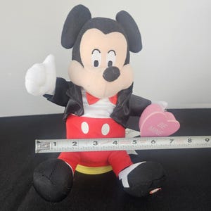 May include: A plush Mickey Mouse figure wearing a black jacket, red trousers with white buttons, and a red bowtie. The figure holds a pink heart-shaped object with the text BE MINE. A metal measuring tape shows the figure is approximately 15 cm wide.