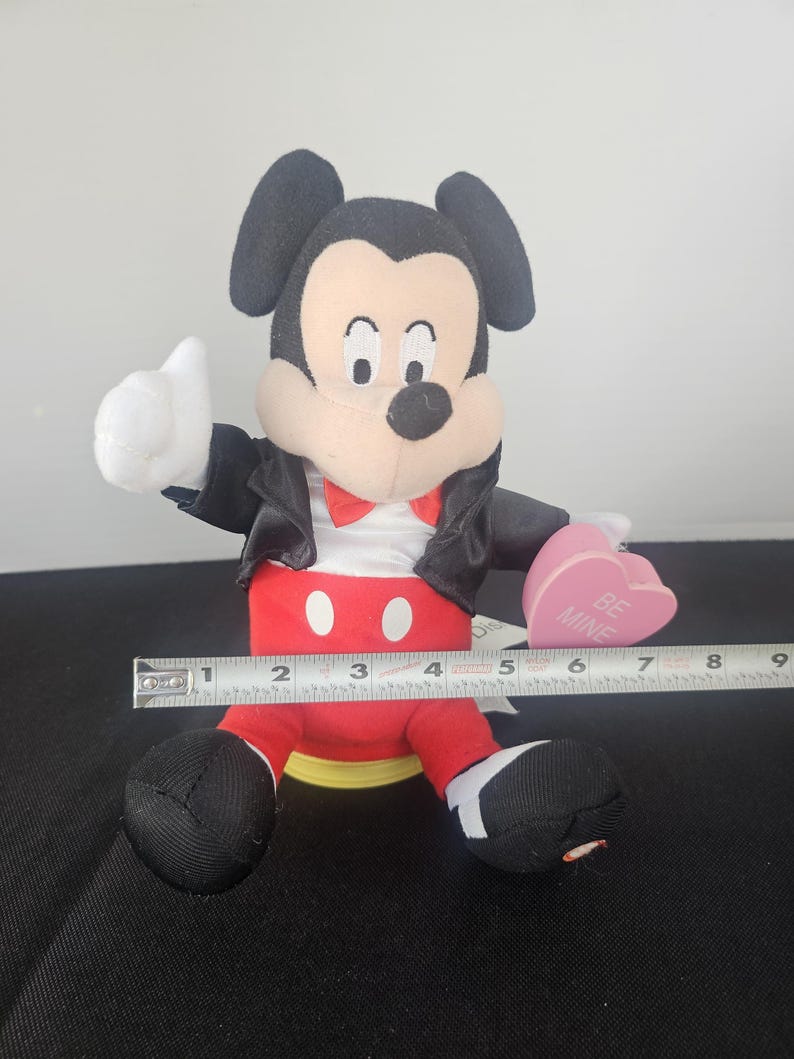 May include: A plush Mickey Mouse figure wearing a black jacket, red trousers with white buttons, and a red bowtie. The figure holds a pink heart-shaped object with the text BE MINE. A metal measuring tape shows the figure is approximately 15 cm wide.