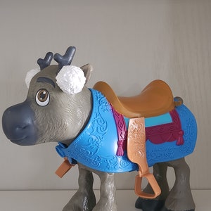 Pu&ograve; includere: Un giocattolo a forma di renna con una sella marrone e una coperta decorativa blu. La renna ha corna grigie, paraorecchie bianchi e zampe marroni. La sella ha staffe marroni e un design decorativo.