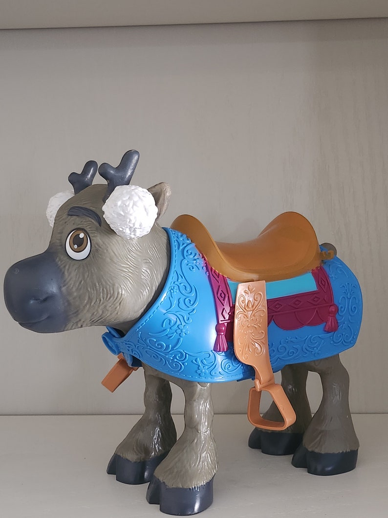 Pu&ograve; includere: Un giocattolo a forma di renna con una sella marrone e una coperta decorativa blu. La renna ha corna grigie, paraorecchie bianchi e zampe marroni. La sella ha staffe marroni e un design decorativo.