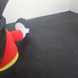 May include: A plush toy featuring a black and white foot with a rounded toe, attached to a red leg. The toy is wearing a black garment and is positioned on a yellow circular base, showcasing a classic cartoon character aesthetic and design.