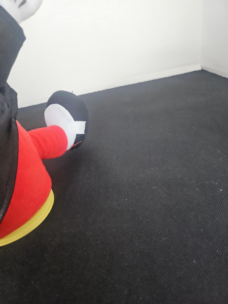 May include: A plush toy featuring a black and white foot with a rounded toe, attached to a red leg. The toy is wearing a black garment and is positioned on a yellow circular base, showcasing a classic cartoon character aesthetic and design.