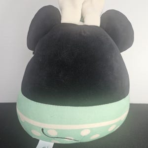 May include: A plush toy shaped like a character's head, featuring large black ears and a black head. The lower portion is mint green with white polka dots and a white stripe. White ears and a tag with the Disney logo are visible.