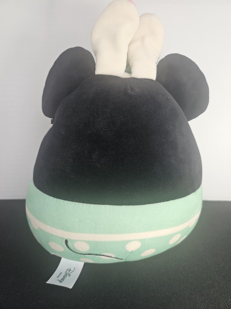 May include: A plush toy shaped like a character's head, featuring large black ears and a black head. The lower portion is mint green with white polka dots and a white stripe. White ears and a tag with the Disney logo are visible.