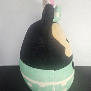 May include: A plush toy shaped like a cartoon character. The toy has black ears, a black head, and a tan face. It wears a mint green skirt with white polka dots and a matching bow. The bow has white polka dots and is mint green.