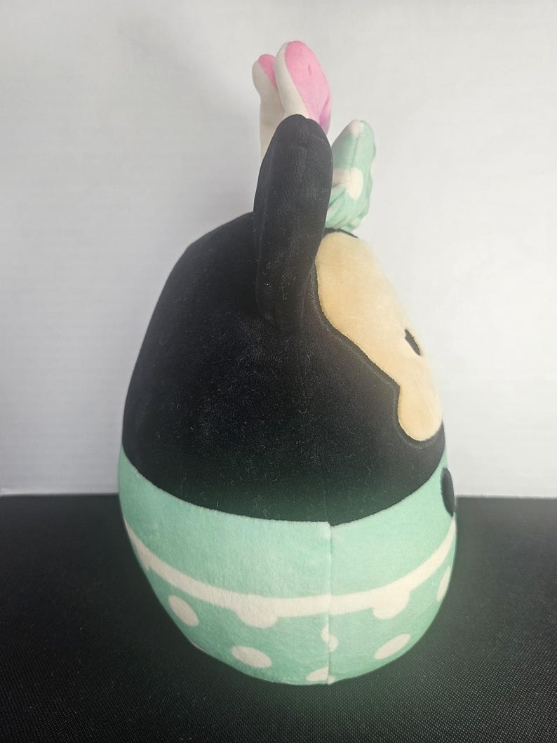 May include: A plush toy shaped like a cartoon character. The toy has black ears, a black head, and a tan face. It wears a mint green skirt with white polka dots and a matching bow. The bow has white polka dots and is mint green.