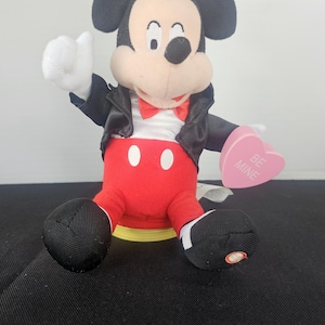 May include: A plush Mickey Mouse figure wearing a black jacket, white shirt, red bowtie, and red trousers with two white buttons. The figure holds a pink heart-shaped object with the text BE MINE printed in white. The figure has black ears and white gloves.