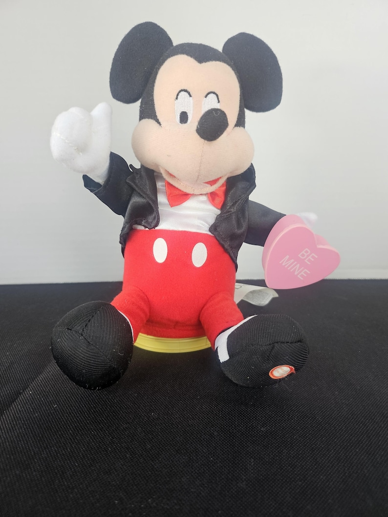 May include: A plush Mickey Mouse figure wearing a black jacket, white shirt, red bowtie, and red trousers with two white buttons. The figure holds a pink heart-shaped object with the text BE MINE printed in white. The figure has black ears and white gloves.