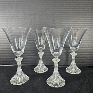 May include: Four clear crystal wine glasses with a flared bowl and ornate stem. The glasses are arranged in a row, showcasing their elegant design. The stems feature a detailed, textured pattern.