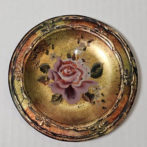 May include: A small, round decorative dish with a gold-colored interior and a detailed, ornate rim. The center features a pink rose with green leaves and small white flowers. The rim has a bronze and green finish.