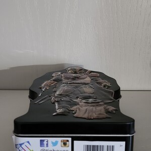Puede incluir: Una caja de metal negra con una imagen 3D de un personaje de Mandalorian de la franquicia Star Wars. El Mandaloriano sostiene un arma y est&aacute; de pie sobre una peque&ntilde;a criatura verde. La caja tiene una etiqueta blanca con el texto "@tinboxco" y "www.tinboxco.com". La etiqueta tambi&eacute;n incluye el texto "Farmingdale, NY 11735", "TIN MADE IN CHINA" y "FABRIQUE EN CHINE".