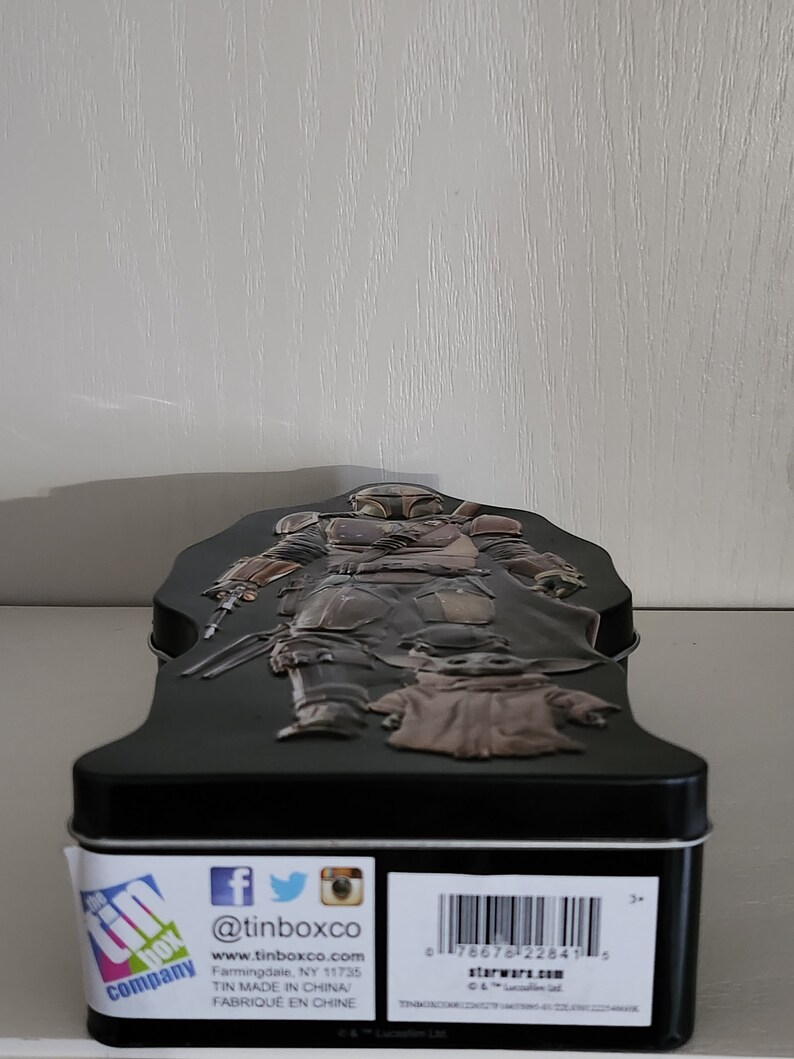 Puede incluir: Una caja de metal negra con una imagen 3D de un personaje de Mandalorian de la franquicia Star Wars. El Mandaloriano sostiene un arma y est&aacute; de pie sobre una peque&ntilde;a criatura verde. La caja tiene una etiqueta blanca con el texto "@tinboxco" y "www.tinboxco.com". La etiqueta tambi&eacute;n incluye el texto "Farmingdale, NY 11735", "TIN MADE IN CHINA" y "FABRIQUE EN CHINE".