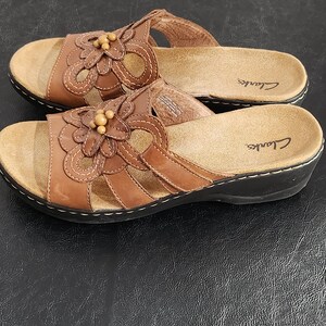 May include: A pair of brown leather slide sandals featuring a floral cutout design on the upper strap, accented with small wooden beads in the center. The sandals have a tan cushioned footbed with the Clarks logo and a black sole with white stitching.