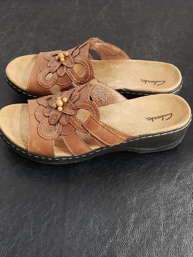May include: A pair of brown leather slide sandals featuring a floral cutout design on the upper strap, accented with small wooden beads in the center. The sandals have a tan cushioned footbed with the Clarks logo and a black sole with white stitching.