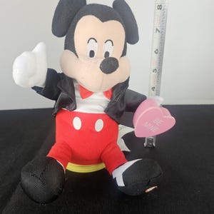 May include: A plush Mickey Mouse figure wearing a black jacket, red trousers, and a red bow tie, holding a pink heart-shaped object with the text BE MINE. A vertical measuring tape next to the figure shows a height of approximately 23 cm.