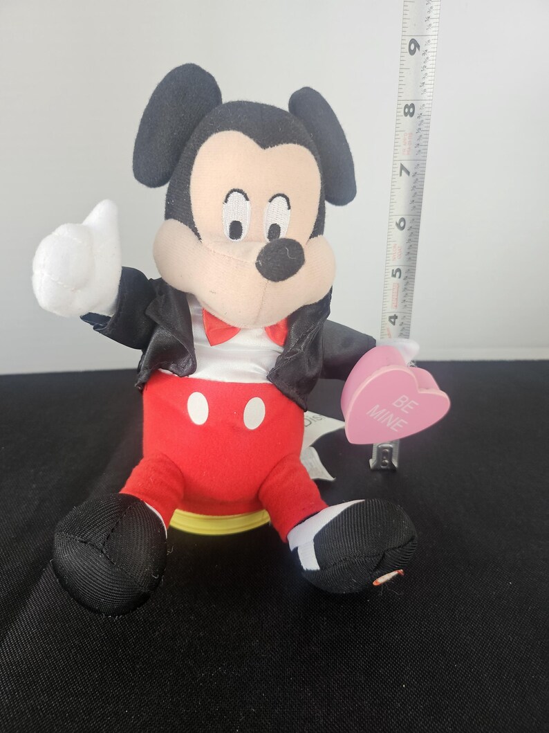May include: A plush Mickey Mouse figure wearing a black jacket, red trousers, and a red bow tie, holding a pink heart-shaped object with the text BE MINE. A vertical measuring tape next to the figure shows a height of approximately 23 cm.
