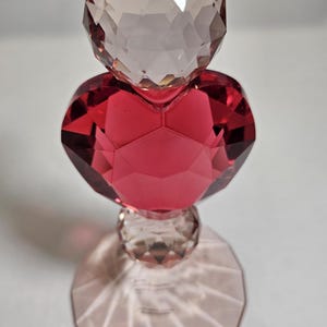Op de afbeelding: Een decoratief kristallen object met een facetontwerp. Het object heeft een heldere, ronde bovenkant, een rood hartvormig midden en een heldere, ronde basis. De basis werpt een schaduw op het witte oppervlak. Het object is waarschijnlijk een decoratief item.