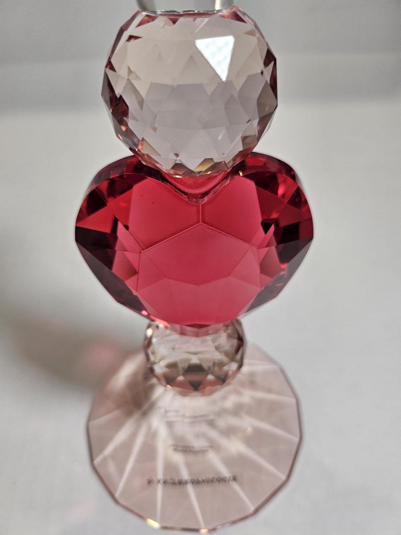 Op de afbeelding: Een decoratief kristallen object met een facetontwerp. Het object heeft een heldere, ronde bovenkant, een rood hartvormig midden en een heldere, ronde basis. De basis werpt een schaduw op het witte oppervlak. Het object is waarschijnlijk een decoratief item.