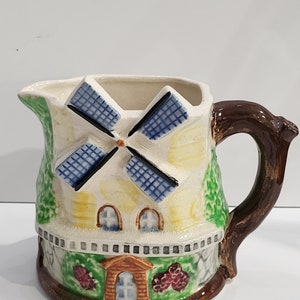 May include: A ceramic pitcher shaped like a windmill with a brown handle. The windmill is painted with blue and white blades, yellow and white walls, and green grass. The base of the pitcher has a brown house with a red roof and a white door.