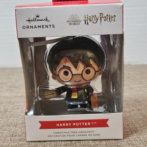 May include: A red and white box with a Harry Potter Christmas tree ornament inside. The ornament is a cartoon depiction of Harry Potter holding a ticket to Platform 9 3/4. The box has the Hallmark and Wizarding World logos.