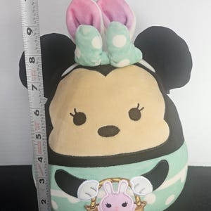 May include: A plush Minnie Mouse figure, approximately 10 inches tall, with black ears, a light tan face, and a mint green and white polka dot outfit. The figure has pink bunny ears and a basket with a pink bunny.