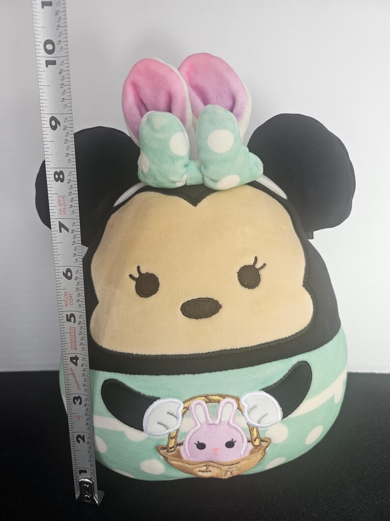 May include: A plush Minnie Mouse figure, approximately 10 inches tall, with black ears, a light tan face, and a mint green and white polka dot outfit. The figure has pink bunny ears and a basket with a pink bunny.