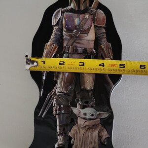 Puede incluir: Una lata de metal negra con un personaje mandaloriano de la franquicia Star Wars. El Mandaloriano lleva un casco y una armadura y sostiene un arma. Un peque&ntilde;o personaje verde, conocido como Grogu, est&aacute; de pie frente al Mandaloriano. La lata mide aproximadamente 15 cm de alto.