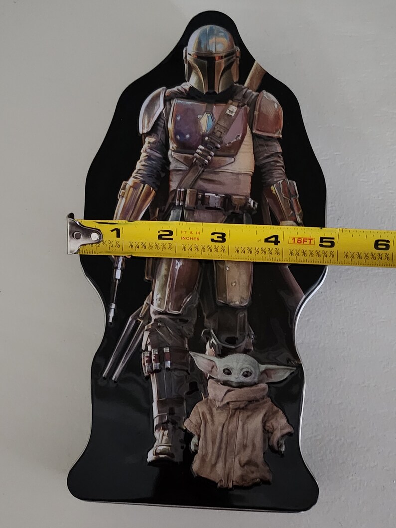 Puede incluir: Una lata de metal negra con un personaje mandaloriano de la franquicia Star Wars. El Mandaloriano lleva un casco y una armadura y sostiene un arma. Un peque&ntilde;o personaje verde, conocido como Grogu, est&aacute; de pie frente al Mandaloriano. La lata mide aproximadamente 15 cm de alto.
