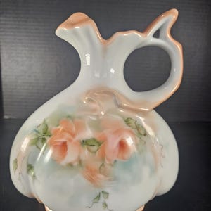 May include: A white ceramic pitcher with a peach-colored rim and handle. The pitcher is decorated with hand-painted peach-colored roses and green leaves. The pitcher has a unique shape with a curved handle and a spout.